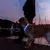 Chien portant un collier lumineux bleu à LED, en promenade au crépuscule près d’un plan d’eau avec des bateaux amarrés en arrière-plan.