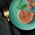 Close-up of wet food on a green plate, garnished with parsley and small cubes. Golden fork next to it.