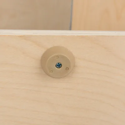 Nahaufnahme einer beige Kunststoffhalterung mit Schraube, befestigt an hellem Holz. Detailansicht eines Möbelstücks oder Zubehörs.
