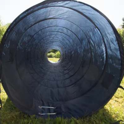 Túnel de entrenamiento para perros de material textil oscuro, colocado sobre césped al aire libre. Vista desde el interior mostrando la estructura circular y los refuerzos metálicos.