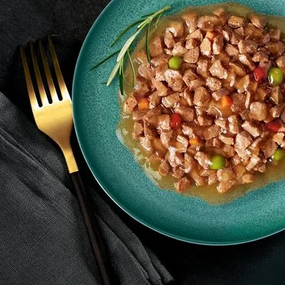 Bord met natvoer in stukjes en gelei, zichtbaar met erwten en wortelstukjes, geserveerd op een groen bord met goudkleurige vork en takje rozemarijn.