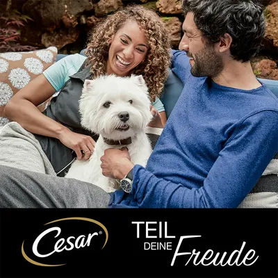 Cesar. Teil deine Freude. Hund sitzt zwischen zwei Personen auf einer Couch.
