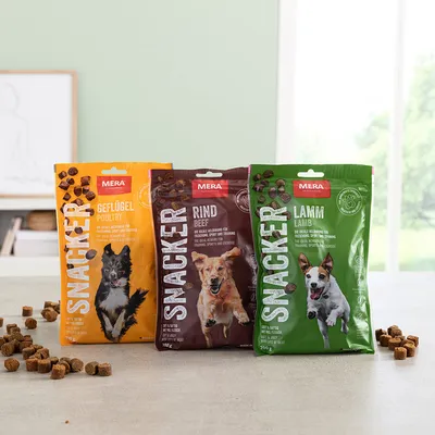 Kolme MERA SNACKER -koiranherkkupussia: Geflügel/Poultry (keltainen), Rind/Beef (ruskea), Lamm/Lamb (vihreä). Pusseissa koirakuvat ja herkkupalat näkyvissä pöydällä.