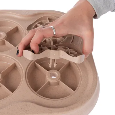 Hand lyfter del av beige aktivitetsleksak i plast med flera runda fack och snurrande delar, avsedd för husdjur.
