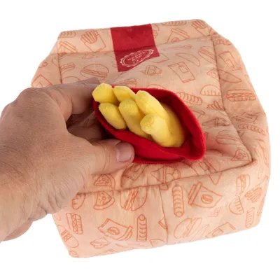 Mână ține o jucărie de pluș în formă de pachet de cartofi prăjiți cu cartofi galbeni din material textil, pe o pernă portocalie cu motive fast-food.