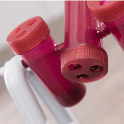 Dettaglio di tubi in plastica rosa trasparente con tappi forati, montati su struttura bianca. Alcuni fori mostrano piccoli oggetti all'interno dei tubi.