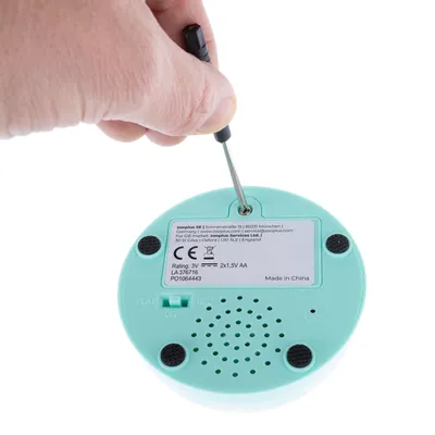 Instrucțiuni pentru schimbarea bateriei: o șurubelniță deschide compartimentul de pe partea inferioară a unui dispozitiv. Specificații: 3V, 2x1,5V AA, Made in China, Zooplus SE, München.