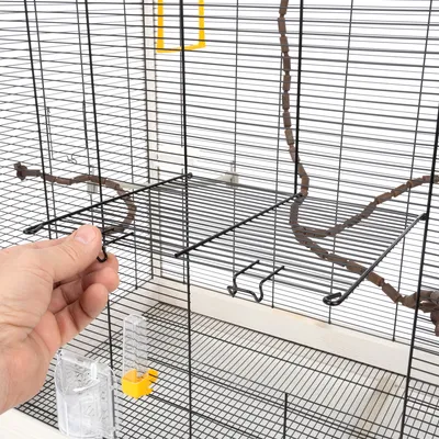 Main ouvrant une porte sur cage en métal noir avec perchoir, abreuvoir transparent à base jaune et mangeoire transparente fixés aux barreaux.