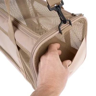 Sac de transport beige pour animaux avec panneaux en maille, main ouvrant une poche latérale zippée, sangle réglable et mousqueton en plastique visibles.