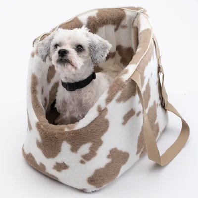 Sac de transport en tissu à motif tacheté beige et blanc, avec un petit chien blanc assis à l’intérieur, harnais noir visible, anse beige sur le côté.