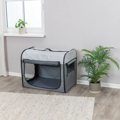 Graue Transportbox mit Netzfenstern und Reißverschluss, geeignet für Innenräume. Steht neben einer Zimmerpflanze und einem Fenster auf Holzboden.