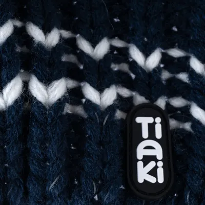 Nahaufnahme eines dunkelblauen Strickmaterials mit weissen Mustern und einem schwarzen Label mit der Aufschrift 'Tiaki'.