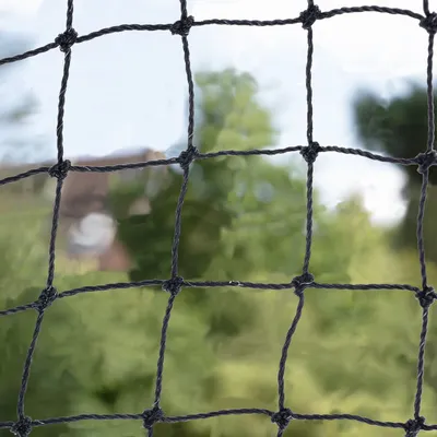 Black knotted netting with square mesh pattern, shown outdoors against a blurred green background. Material appears to be twisted synthetic fibre.