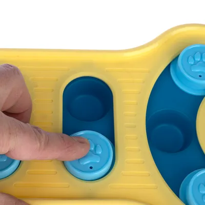 Main appuyant sur un bouton bleu en forme de patte sur un jouet interactif jaune et bleu pour animaux, éléments en plastique visibles.