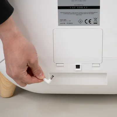Hand showing connection of a mains plug to the automatic cat toilet. Product details and CE marking visible on the back.