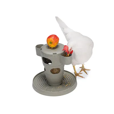 Comedero gris con varias aberturas, lleno de granos y una manzana encima. Gallina blanca come de una de las aberturas.