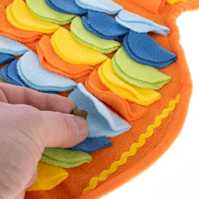 Ręka wkłada smakołyk do kolorowych materiałowych kieszeni interaktywnej zabawki. Wspiera aktywności węchowe i poszukiwawcze dla zwierząt.