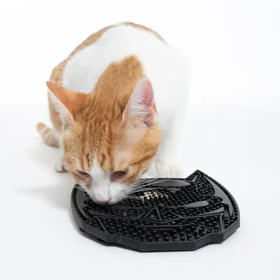 Röd och vit katt slickar mat från en svart, strukturerad matta i form av ett katt huvud.