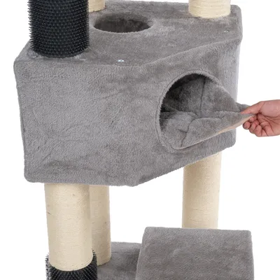 Arbre à chat gris avec niche en tissu, coussin amovible visible tenu par une main, poteaux recouverts de sisal et brosse noire fixée sur un montant.