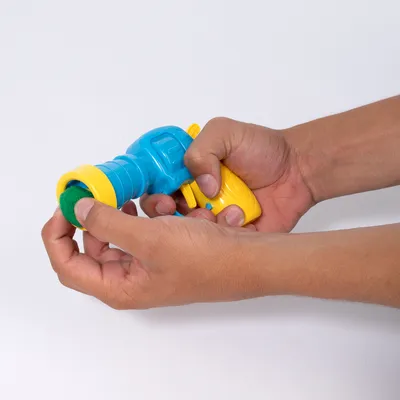 Hände setzen einen grünen Ball in eine gelb-blaue Spielzeugpistole ein, demonstrieren die Verwendung des Produkts.