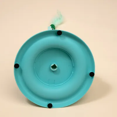 Vue du dessous d’un jouet pour chat en plastique turquoise, avec quatre patins antidérapants noirs et une plume verte fixée sur le bord.
