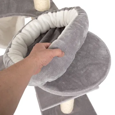 Detalle de una mano sujetando una cama redonda acolchada gris y blanca de tela, parte de un árbol rascador para gatos con postes forrados de cuerda.