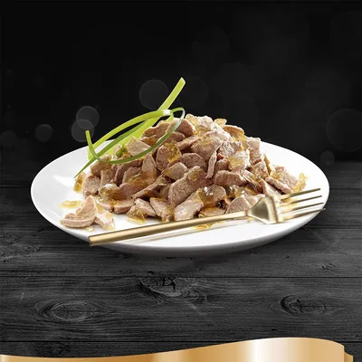 Hvid tallerken med stykker af vådfoder i gelé, pyntet med grønne strimler. Guldgaffel og -kniv ved siden af på sort træbord. Ingen synlige mærker eller tekst.