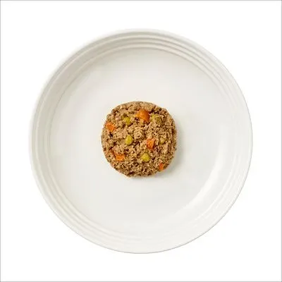 Portie natvoer met zichtbare stukjes wortel en erwt op een wit bord, bovenaanzicht. Geen merknaam of verpakking zichtbaar.