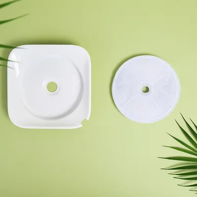 Recambio de filtro redondo blanco y base cuadrada de plástico blanco para fuente de agua, ambos con orificio central, sobre fondo verde.