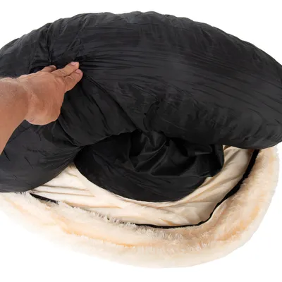 Hand lifting the black cover of a round, plush pet bed to show removable or washable inner padding.