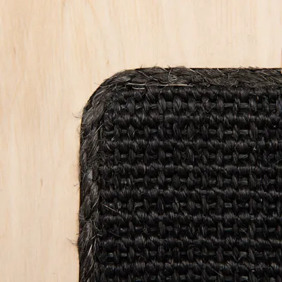 Detail černé tkané podložky z textilu na světlém dřevěném povrchu, viditelná struktura materiálu a zaoblený roh.