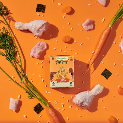Naturediet Feel Good Turkey & Chicken food pack surrounded by raw chicken, carrots, rice grains and vegetable pieces, highlighting main ingredients.
