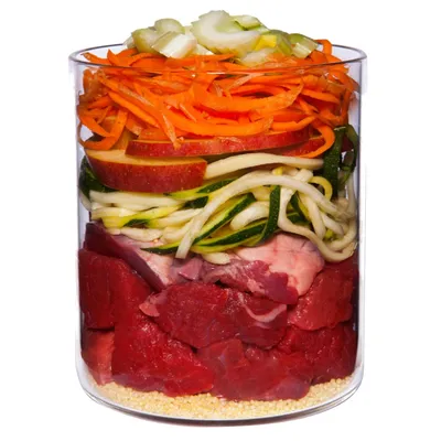Glass container with visible layers of raw beef chunks, millet, courgette strips, apple slices, grated carrot and chopped celery. All ingredients clearly separated in the jar.