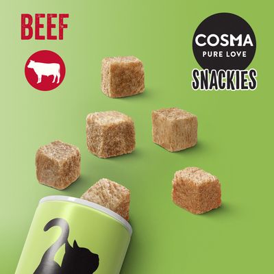 Cubos de snack para gatos saliendo de un envase verde con silueta de gato. Texto visible: BEEF, COSMA PURE LOVE SNACKIES. Icono de vaca blanca en círculo rojo.