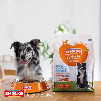 Smølke Adult Medium Complete all-in-one hondenvoer, Daily Balance, uniek eiwitprofiel. Hond naast voerbak en voederzak. Slogan: feed the love.