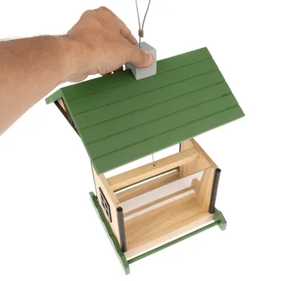 Mangeoire à oiseaux en bois avec toit vert, suspendue par un câble métallique, main tenant le crochet de suspension. Parois latérales transparentes visibles.