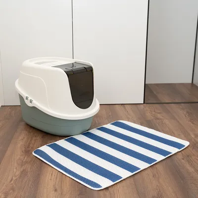 Geschlossene Katzentoilette mit weissem Deckel und schwarzer Klappe, daneben gestreifte Matte in Blau und Weiss auf Holzboden.