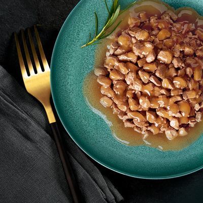 Bord met stukjes natvoer in saus, geserveerd op een groen bord met goudkleurige vork en zwarte servet. Geen merknaam of productinformatie zichtbaar.