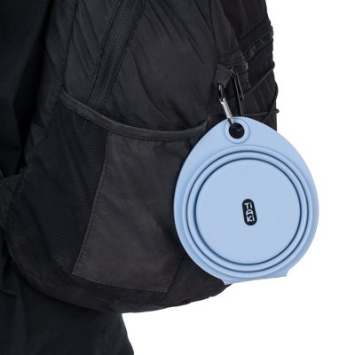 Hopfällbar, blå Tiaki-reseskål i silikon, fäst vid en svart ryggsäck med karbinhake.