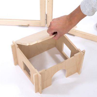 Maison en bois clair pour petits animaux, toit amovible tenu par une main, fenêtres découpées sur les côtés et entrée en arc visible à l'avant.