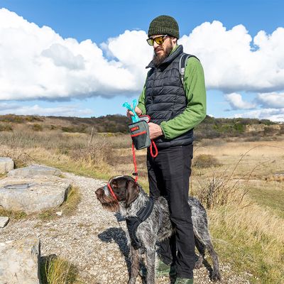 Persoon houdt een hondenriem en een tas met hondenpoepzakjes, hond draagt een tuigje en muilkorf in een landschap.