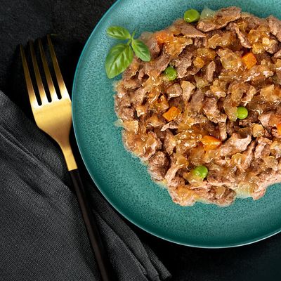 Assiette contenant de la nourriture humide en morceaux avec sauce, petits pois et carottes visibles, présentée sur une assiette verte à côté d'une fourchette dorée.