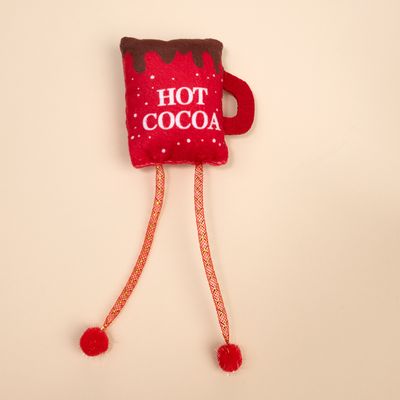 Червена плюшена играчка във формата на чаша с надпис 'Hot Cocoa', два дълги шнура и червени помпони накрая.