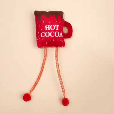 Червена плюшена играчка във формата на чаша с надпис 'Hot Cocoa', два дълги шнура и червени помпони накрая.