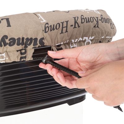 Hände ziehen an einem schwarzen Kordelzug, um einen beige bedruckten Stoffbezug auf einer Transportbox zu befestigen.