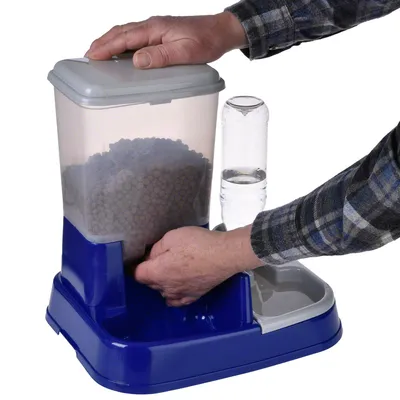 Dispenser de hrană și apă cu bază albastră, recipient transparent pentru hrană uscată și sticlă pentru apă. Mâinile arată umplerea dispenserului.