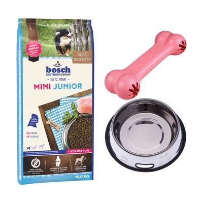Confezione Bosch Medium Junior 15 kg, ciotola in metallo, osso rosa in gomma, simbolo percentuale rosso per offerta. Testo visibile: 'BIS 12. MONAT', 'KOLOSTRUM', '26 PROTEIN', '13 FETTGEHALT'.