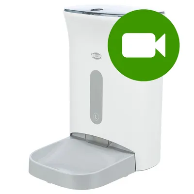 Automatyczny podajnik karmy Trixie, biały z szarą miską, widoczny symbol kamery w zielonym kółku na górze po prawej stronie.