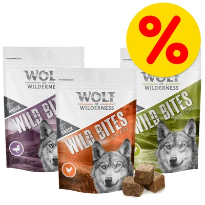 Wolf of Wilderness Wild Bites, tres bolsas de snack sin cereales con texto visible: Grain Free Snack. Variedades Wide Acres y Green Fields. Cubos de snack delante. Símbolo de porcentaje en amarillo.