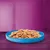 Croquettes ou bouchées pour animaux dans une gamelle bleue posée sur une table violette, sans marque ni texte visible.